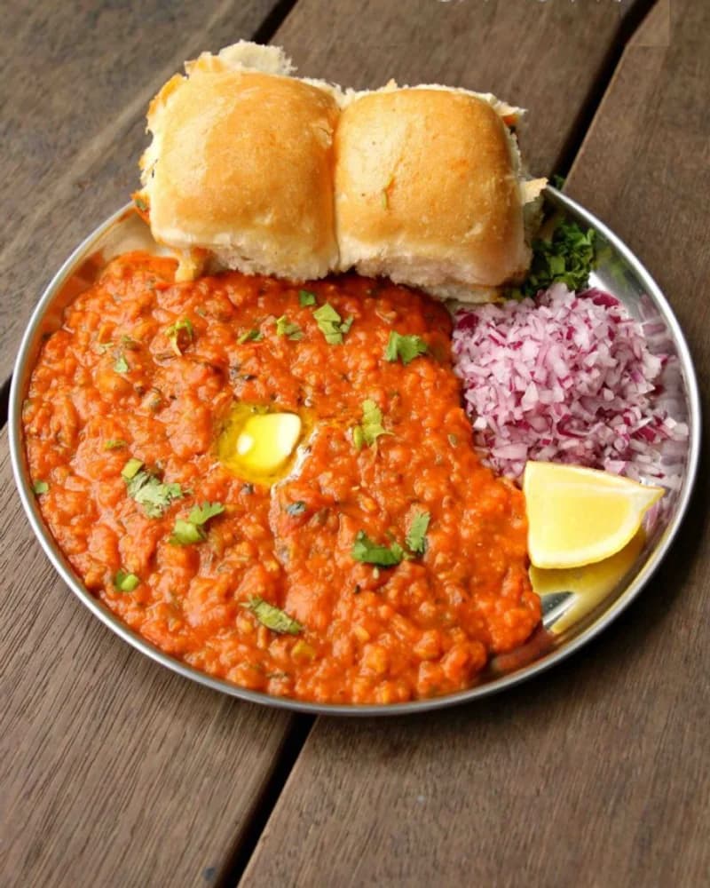 Pav bhaji on a steel plate with butter-toasted pav, chopped onion, coriander and a lemon wedge